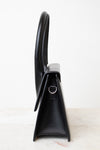 A black leather shoulder bag shown from a side profile view, standing upright on a light marble surface against a white background.