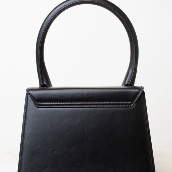 A black leather handbag with a structured, geometric shape and a single integrated top handle, photographed against a clean white background.