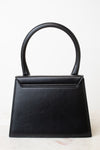 A black leather handbag with a structured, geometric shape and a single integrated top handle, photographed against a clean white background.