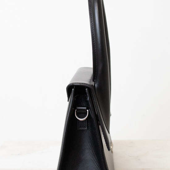 A black leather handbag shown from a side profile view, featuring a structured triangular silhouette, a top handle, and a small silver-tone metal D-ring, set against a plain white background.