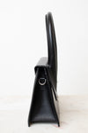 A black leather handbag shown from a side profile view, featuring a structured triangular silhouette, a top handle, and a small silver-tone metal D-ring, set against a plain white background.
