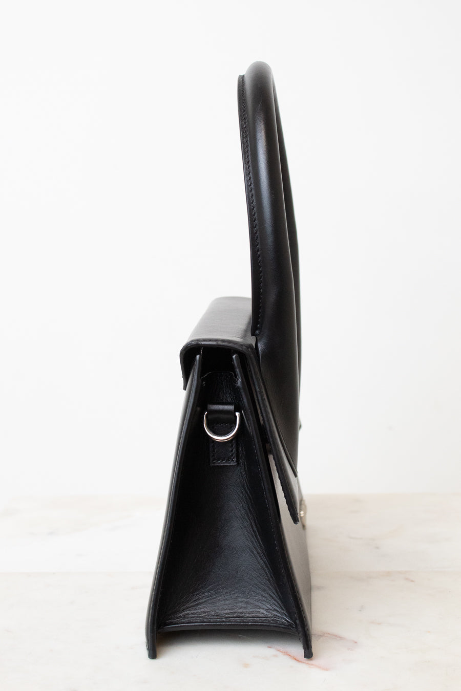 A black leather handbag shown from a side profile view, featuring a structured triangular silhouette, a top handle, and a small silver-tone metal D-ring, set against a plain white background.