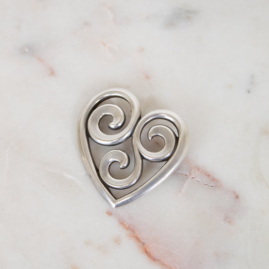 Silver heart-shaped pendant with swirling designs on a marble surface