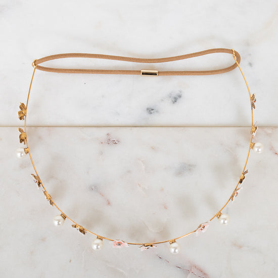 A delicate gold chain headband with small gold leaf charms, white pearl beads, and a tan elastic band, displayed on a white marble surface.