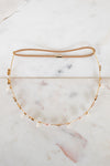 A delicate gold chain headband with small gold leaf charms, white pearl beads, and a tan elastic band, displayed on a white marble surface.