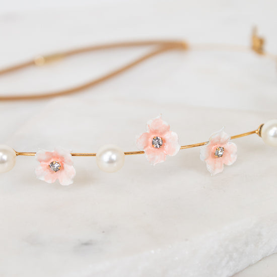 A delicate gold chain headband featuring three light pink flower charms with clear rhinestones and white faux pearls, displayed on a white marble slab.