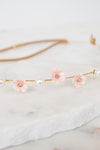 A delicate gold chain headband featuring three light pink flower charms with clear rhinestones and white faux pearls, displayed on a white marble slab.
