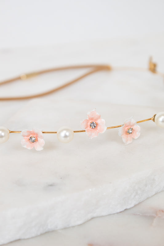 A delicate gold chain headband featuring three light pink flower charms with clear rhinestones and white faux pearls, displayed on a white marble slab.