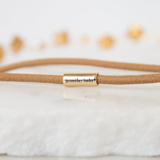 A close-up of a tan cord bracelet with a gold metal cylinder clasp engraved with the name 'jennifer behr' in black script, resting on a white textured stone surface.
