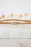 A close-up of a tan cord bracelet with a gold metal cylinder clasp engraved with the name 'jennifer behr' in black script, resting on a white textured stone surface.