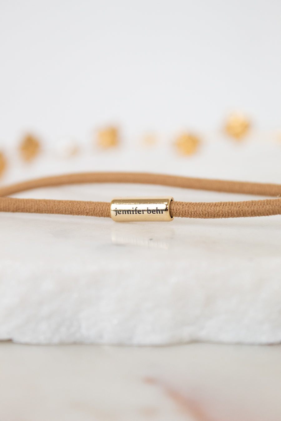 A close-up of a tan cord bracelet with a gold metal cylinder clasp engraved with the name 'jennifer behr' in black script, resting on a white textured stone surface.