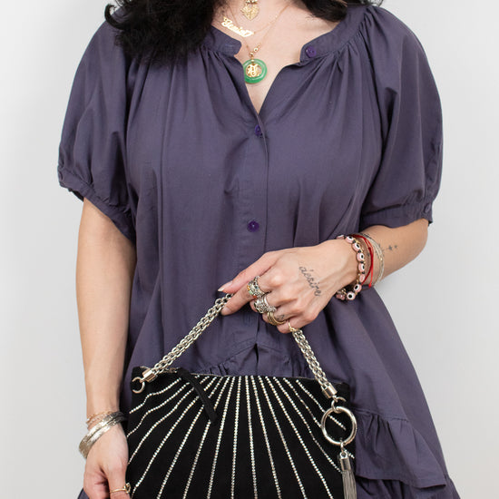 A woman wearing a purple short-sleeve blouse with puff sleeves and a ruffled hem, holding a black  clutch bag with silver chain straps and tassel details.