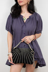 A woman wearing a purple short-sleeve blouse with puff sleeves and a ruffled hem, holding a black  clutch bag with silver chain straps and tassel details.