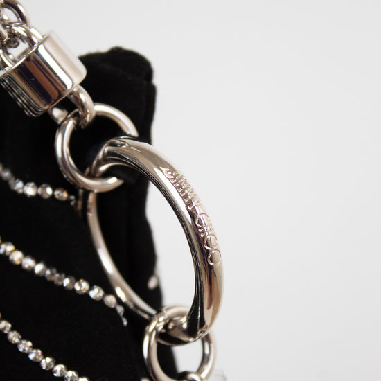 Close-up detail of a black handbag featuring a silver-tone metal chain strap, a large interlocking O-ring, and a padlock charm, with the brand name 'Dior' engraved on the ring.