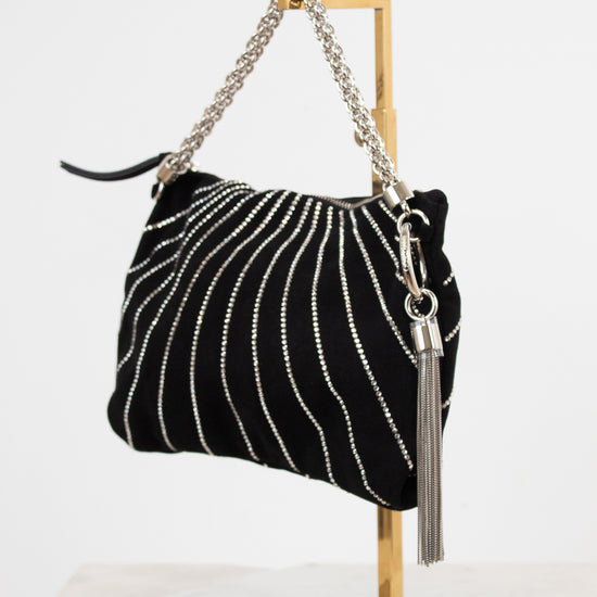 A black velvet shoulder bag with vertical silver rhinestone stripes, a silver chain strap, and a silver tassel charm, displayed on a gold metal stand against a white background.