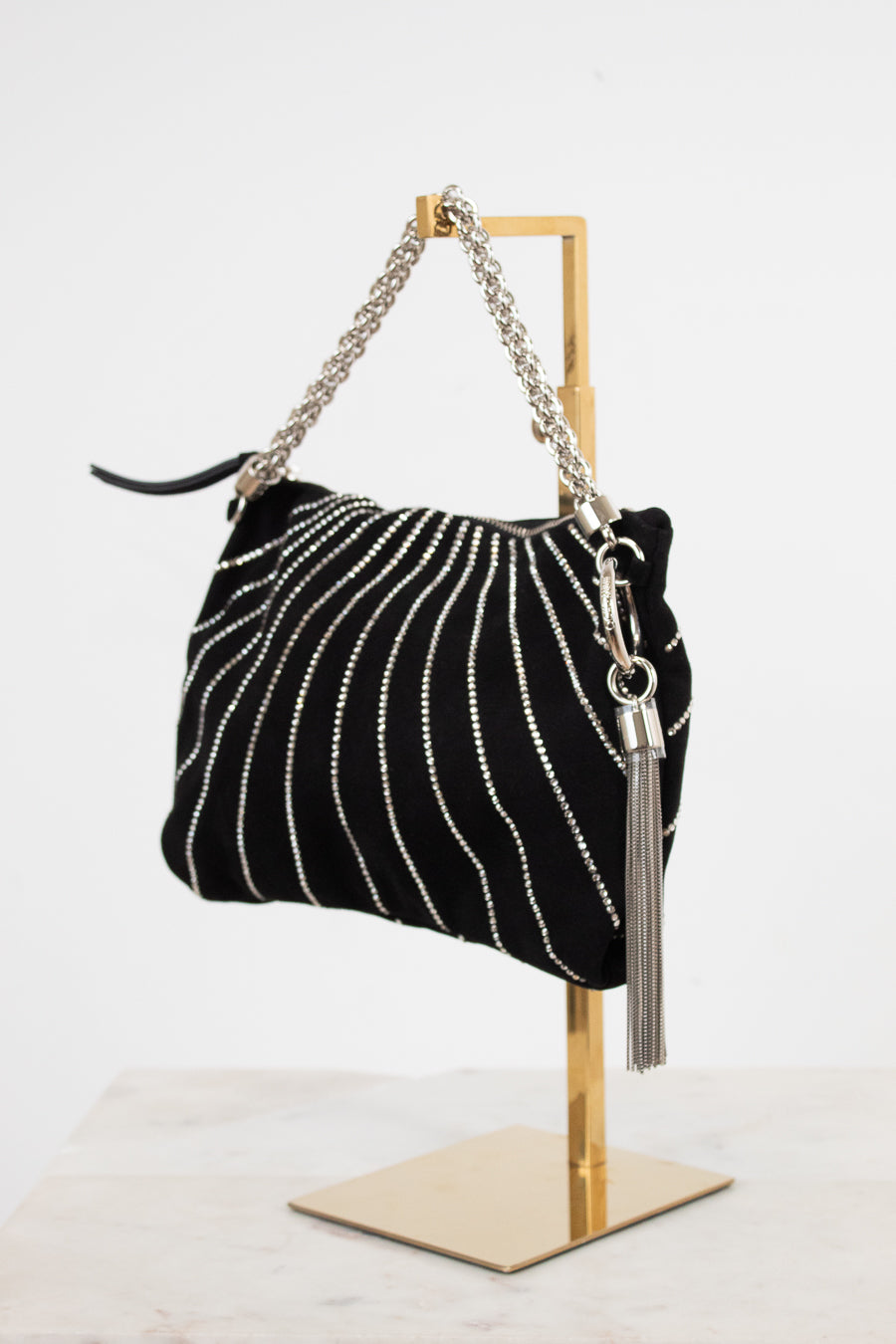 A black velvet shoulder bag with vertical silver rhinestone stripes, a silver chain strap, and a silver tassel charm, displayed on a gold metal stand against a white background.
