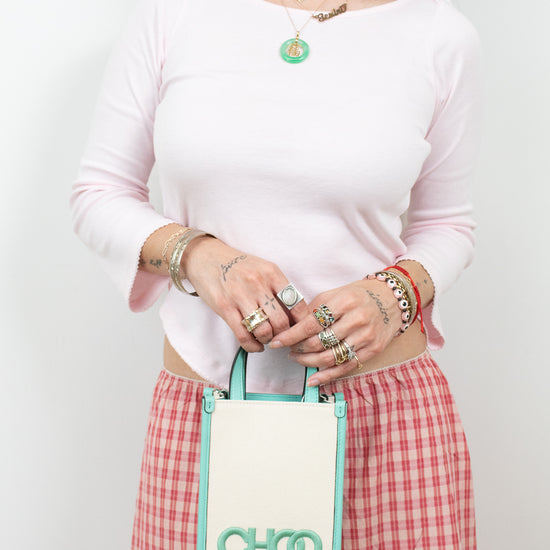 A woman wearing a light pink long-sleeve top and a red-and-white gingham skirt holds a small cream tote bag with mint green trim and the brand name CHOO visible on the front.