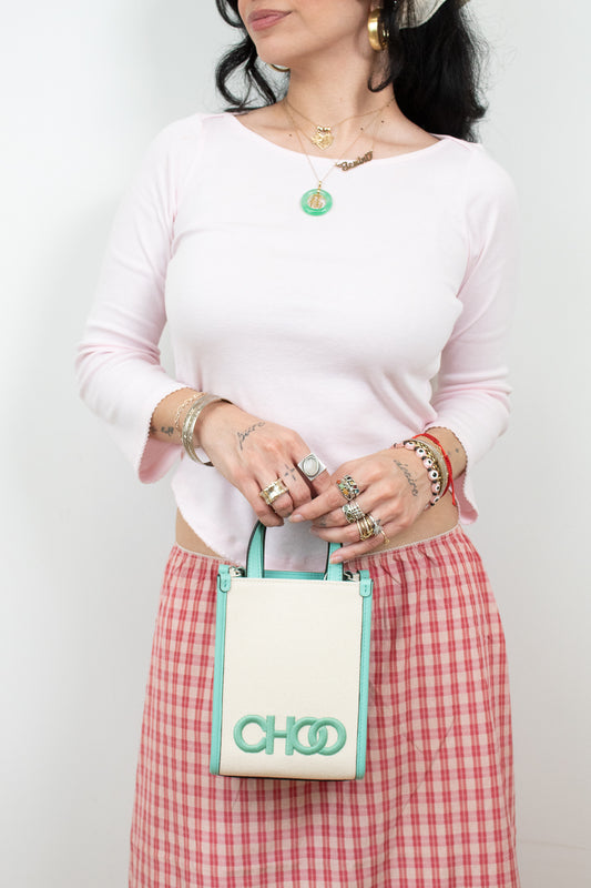 A woman wearing a light pink long-sleeve top and a red-and-white gingham skirt holds a small cream tote bag with mint green trim and the brand name CHOO visible on the front.