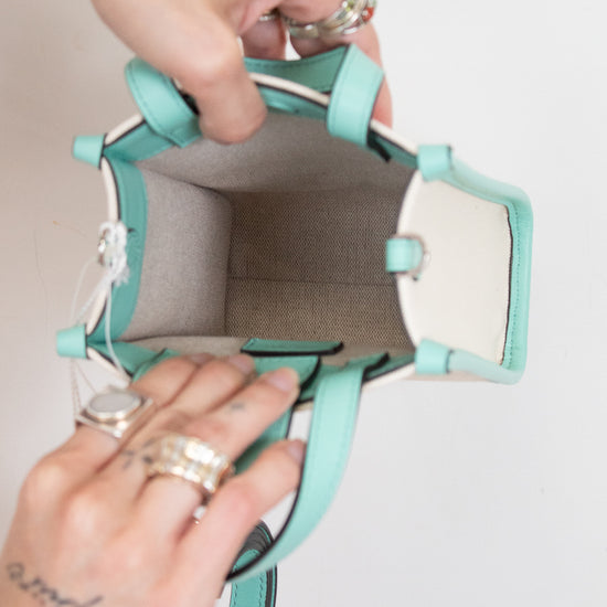 A top-down view of a mint green and white handbag being held open by two hands wearing silver rings, against a plain white background.