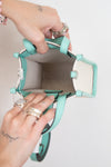 A top-down view of a mint green and white handbag being held open by two hands wearing silver rings, against a plain white background.