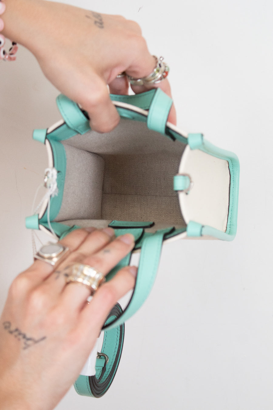 A top-down view of a mint green and white handbag being held open by two hands wearing silver rings, against a plain white background.