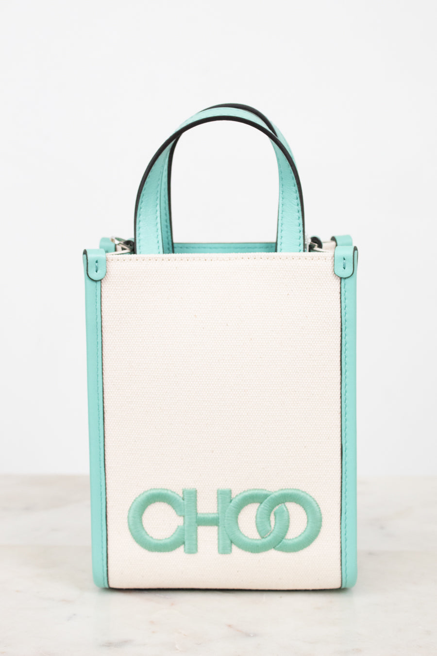 A structured mini tote bag with a cream-colored front panel and mint green trim, handles, and logo lettering, displayed on a light marble surface against a white background.