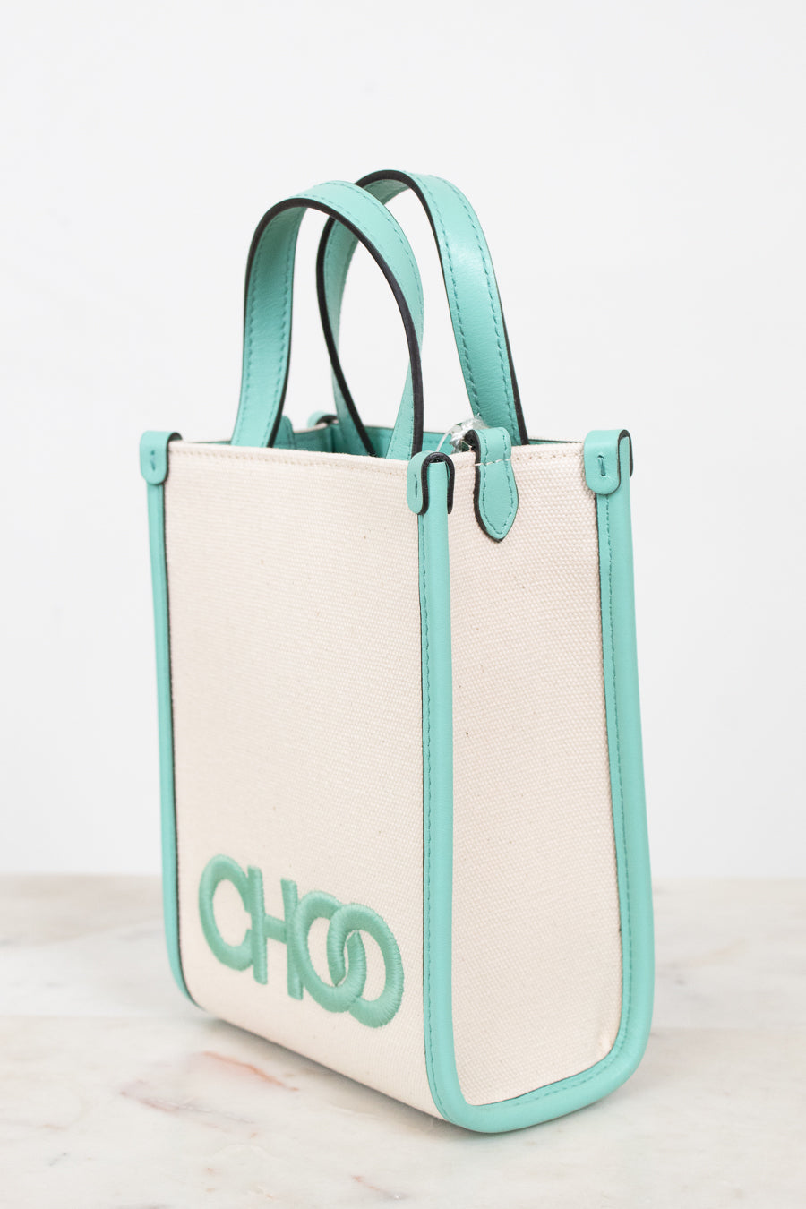 A structured tote bag with a cream canvas body and mint green leather trim, handles, and side borders. The bag features a large mint green 'CHOO' logo on the front, set against a clean white background.