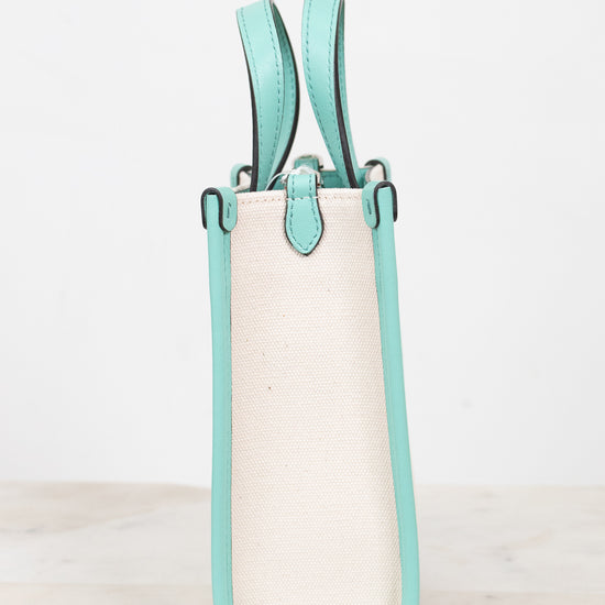 A structured mini tote bag with a cream canvas body and mint green leather trim and handles, standing upright and showing the side profile on a light marble surface against a white background.