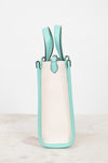 A structured mini tote bag with a cream canvas body and mint green leather trim and handles, standing upright and showing the side profile on a light marble surface against a white background.