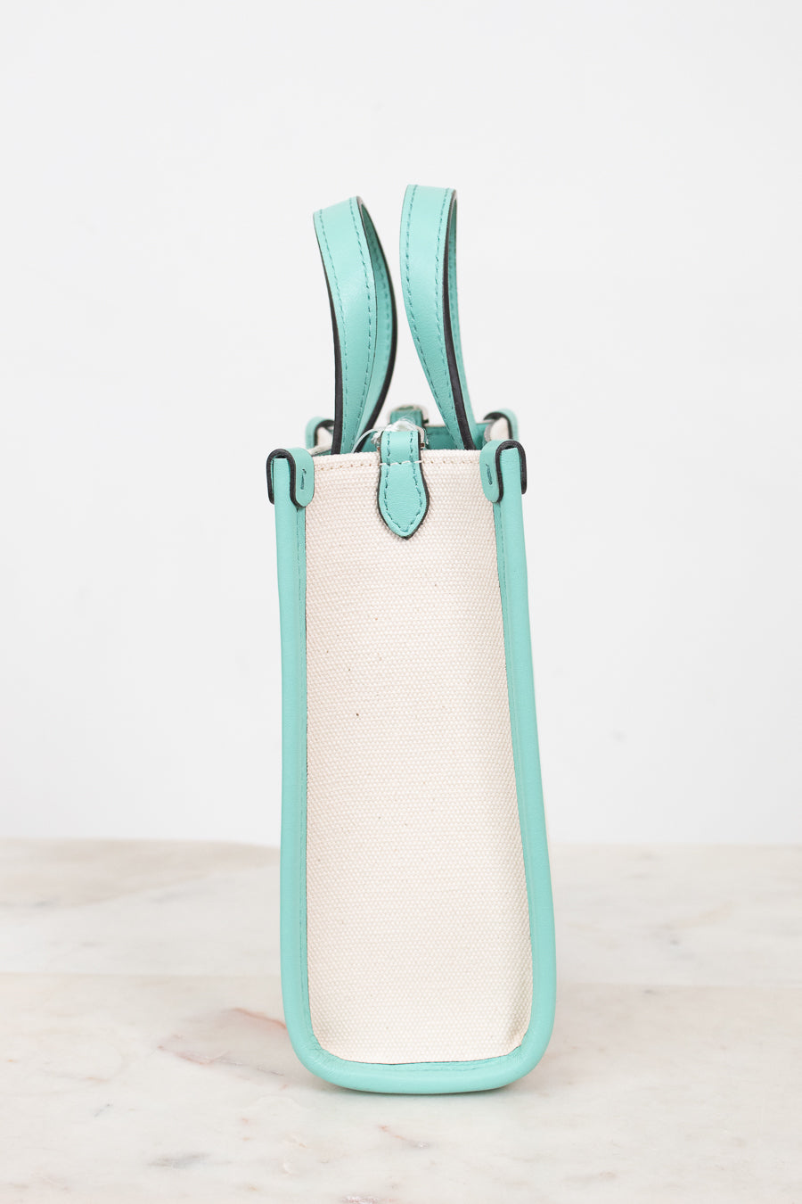 A structured mini tote bag with a cream canvas body and mint green leather trim and handles, standing upright and showing the side profile on a light marble surface against a white background.