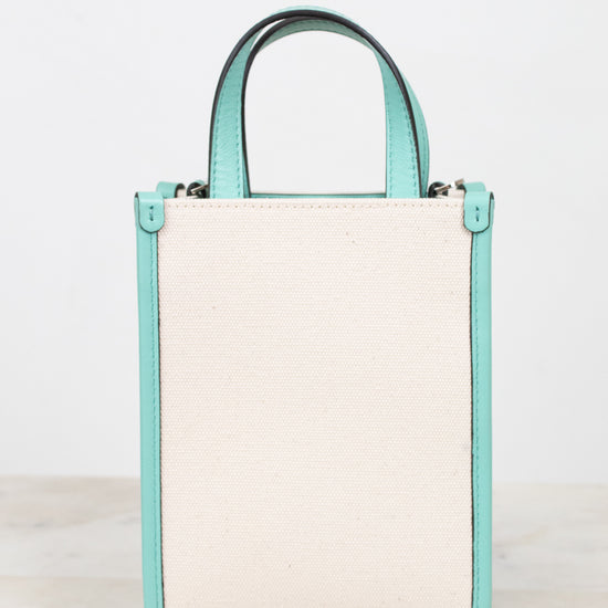 A structured mini tote bag with a large off-white textured back panel and mint green side panels and top handle, shown on a light marble surface against a white background.