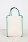 A structured mini tote bag with a large off-white textured back panel and mint green side panels and top handle, shown on a light marble surface against a white background.