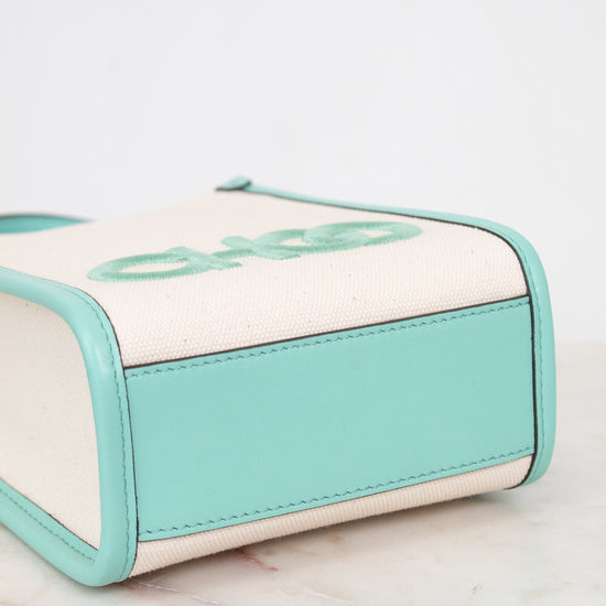 A close-up of a small handbag or pouch with a cream canvas body, mint green leather trim, and a matching mint green embroidered logo on the top, resting on a light marble surface.