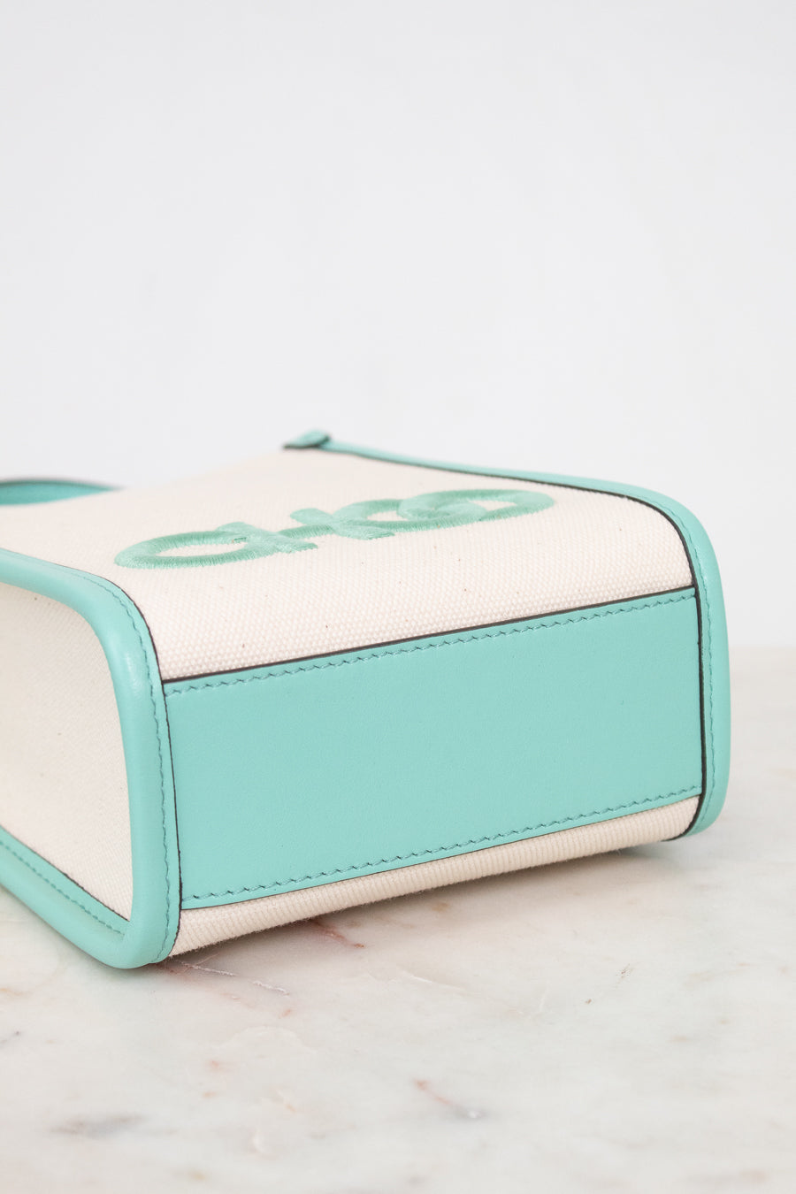 A close-up of a small handbag or pouch with a cream canvas body, mint green leather trim, and a matching mint green embroidered logo on the top, resting on a light marble surface.