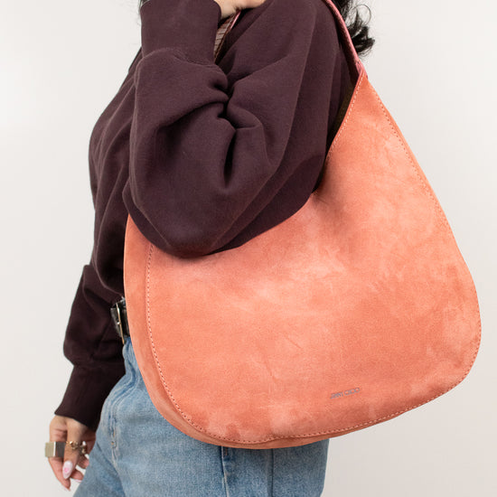 Person wearing a brown sweatshirt and blue jeans with a large pinkish-brown bag over their shoulder against a plain background.