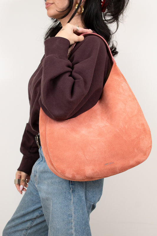 Person wearing a brown sweatshirt and blue jeans with a large pinkish-brown bag over their shoulder against a plain background.