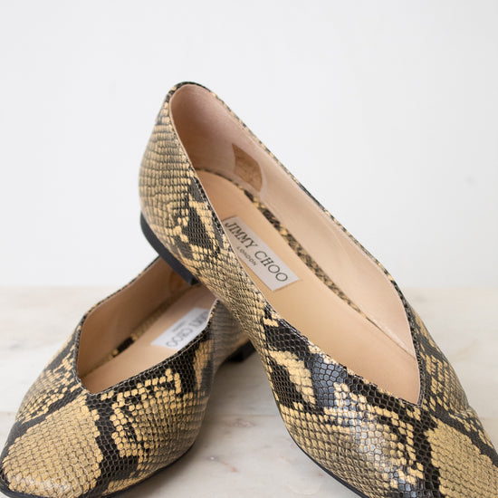 A pair of Jimmy Choo snakeskin pointed-toe flats in beige, black, and brown, displayed on a light marble surface against a white background.