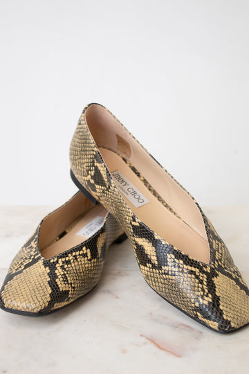 A pair of Jimmy Choo snakeskin pointed-toe flats in beige, black, and brown, displayed on a light marble surface against a white background.