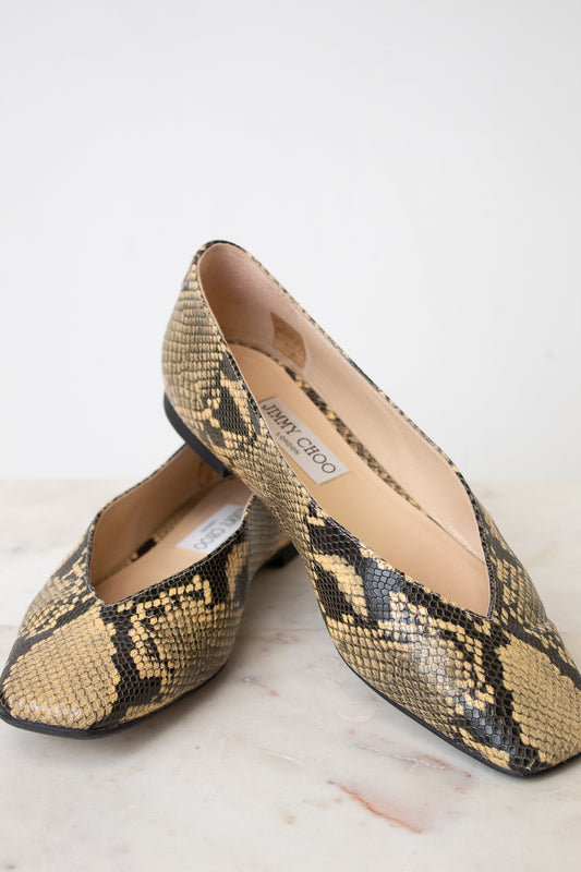A pair of Jimmy Choo snakeskin pointed-toe flats in beige, black, and brown, displayed on a light marble surface against a white background.