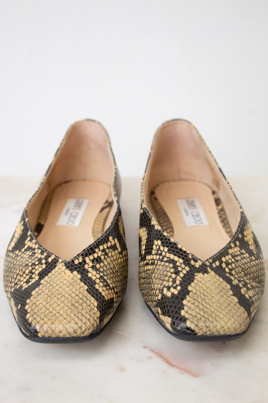 A pair of Jimmy Choo snakeskin ballet flats with a beige, black, and brown python pattern, shown on a light marble surface against a white background.