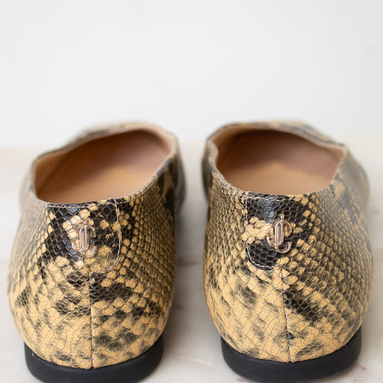 A pair of women's slip-on flats with a snakeskin pattern in tan and black, featuring a small gold 'JC' logo on the heel, photographed from the back on a light marble surface.