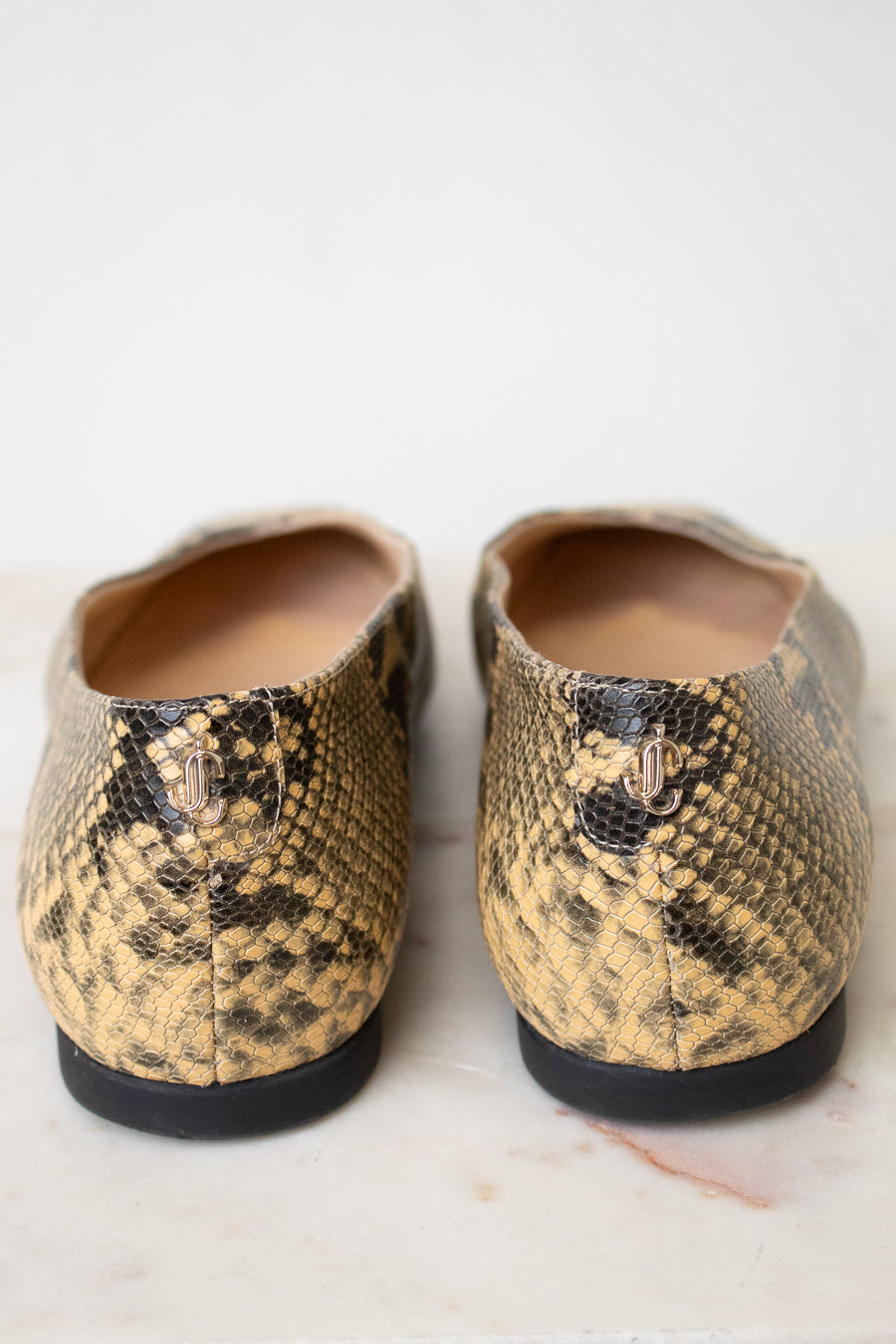 A pair of women's slip-on flats with a snakeskin pattern in tan and black, featuring a small gold 'JC' logo on the heel, photographed from the back on a light marble surface.