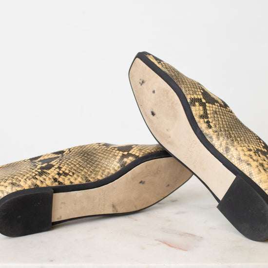 A pair of snakeskin-patterned flats with black soles, shown from the bottom on a white marble surface against a white background.