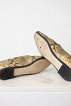 A pair of snakeskin-patterned flats with black soles, shown from the bottom on a white marble surface against a white background.