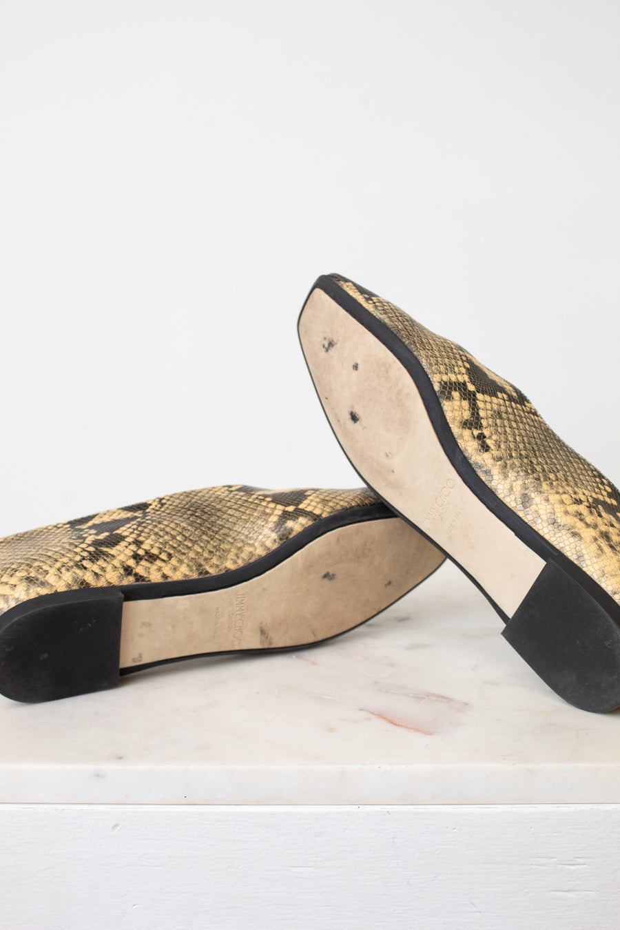 A pair of snakeskin-patterned flats with black soles, shown from the bottom on a white marble surface against a white background.