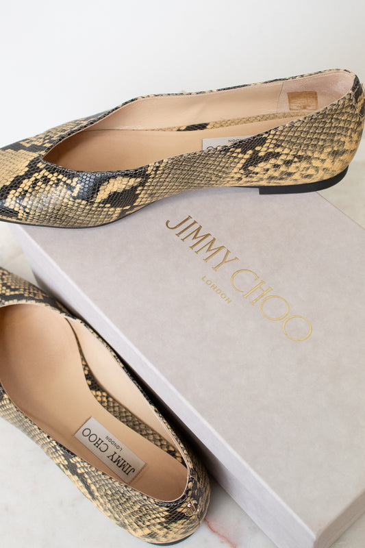 A pair of Jimmy Choo London pointed-toe flat shoes in a snakeskin pattern, displayed on top of and next to their light gray brand box.