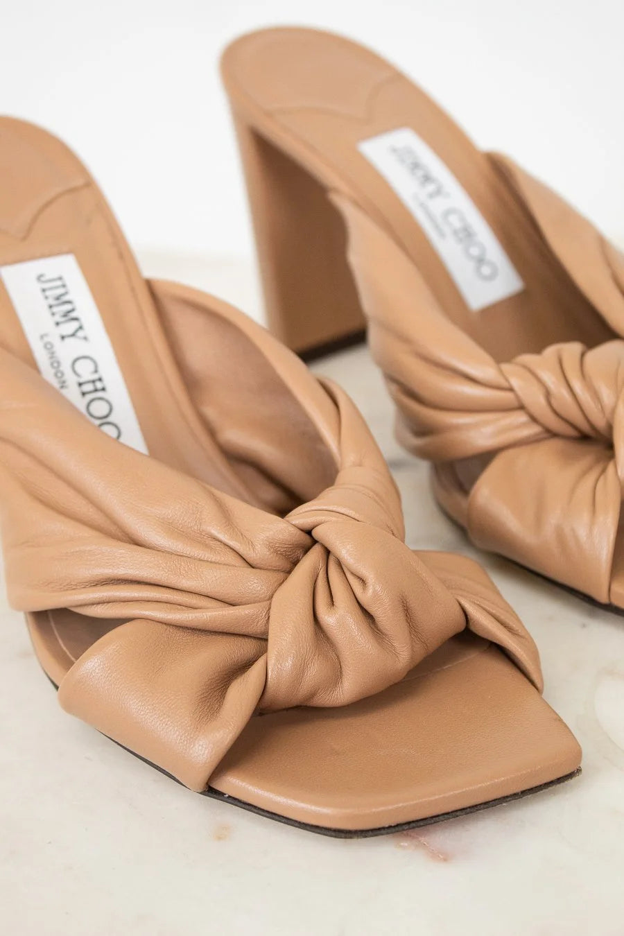 Pair of tan high-heeled shoes with knotted design on a light background, featuring Jimmy Choo branding.