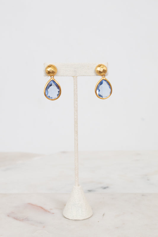 Gold-border earrings with blue teardrop stones on a white stand against a light background