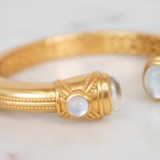 A gold bangle bracelet with ornate detailing and round white moonstone accents, resting on a light marble surface.