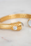 A gold bangle bracelet with ornate detailing and round white moonstone accents, resting on a light marble surface.